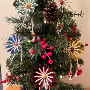 May include: A decorated Christmas tree with various ornaments, including flower-shaped glass ornaments in blue, white, and yellow. The tree also features red berries, pinecones, and white string accents, creating a festive holiday display.
