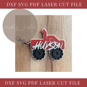 Może przedstawiać: Czerwono-czarny brelok do kluczy w kształcie ciężarówki z napisem "HUDSON" w białym skrypcie. Brelok jest przymocowany do przezroczystego, okrągłego tagu z napisem "Southern Cooklin Designs". Tło to jasne usłojenie drewna.