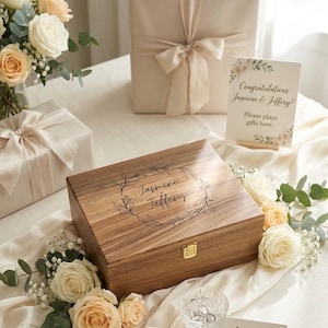 May include: A wooden box with a gold clasp, engraved with "Jasmine & Jeffery" in a floral wreath. The box is surrounded by white and peach roses, with a sign that reads "Congratulations Jasmine & Jeffery! Please place gifts here."