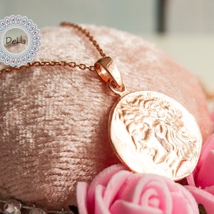 Pode incluir: Um colar com pingente em ouro rosa com um relevo detalhado de uma figura cl&aacute;ssica em uma moeda circular. O pingente pende de uma corrente delicada. O colar &eacute; exibido em uma superf&iacute;cie de veludo rosa, rodeado por rosas cor-de-rosa.