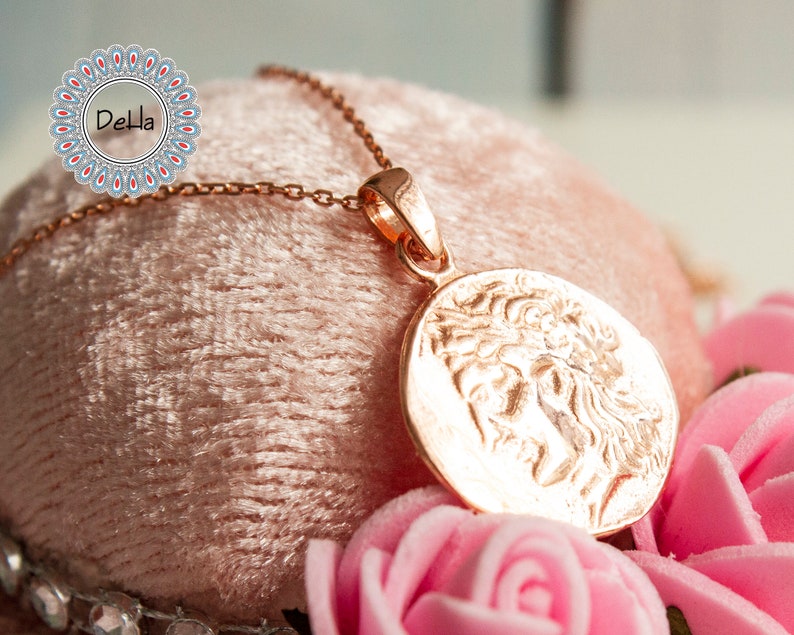 Pode incluir: Um colar com pingente em ouro rosa com um relevo detalhado de uma figura cl&aacute;ssica em uma moeda circular. O pingente pende de uma corrente delicada. O colar &eacute; exibido em uma superf&iacute;cie de veludo rosa, rodeado por rosas cor-de-rosa.