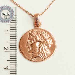 Pode incluir: Colar com pingente em ouro rosa com um berloque em forma de moeda que retrata um perfil com uma coroa de louros. O pingente tem aproximadamente 2,5 cm de di&acirc;metro e pende de uma corrente delicada. O colar &eacute; exibido em uma superf&iacute;cie branca.