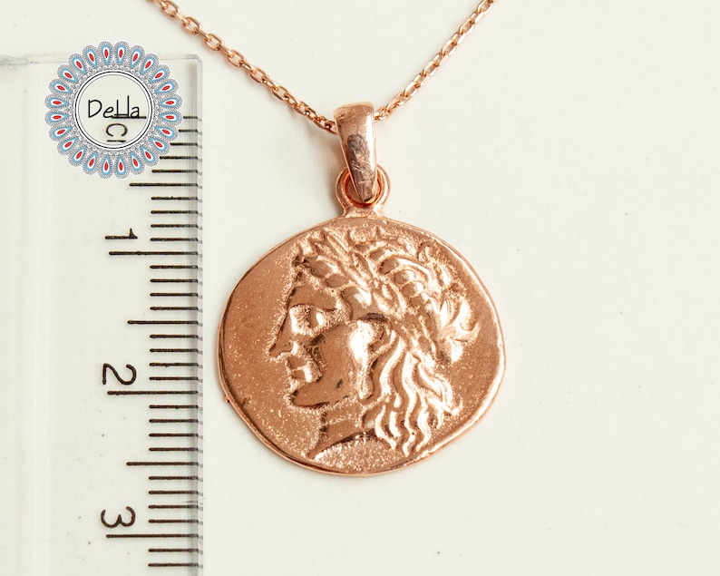 Pode incluir: Colar com pingente em ouro rosa com um berloque em forma de moeda que retrata um perfil com uma coroa de louros. O pingente tem aproximadamente 2,5 cm de di&acirc;metro e pende de uma corrente delicada. O colar &eacute; exibido em uma superf&iacute;cie branca.