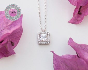 May include: A silver necklace with a square pendant featuring a large, clear square-cut stone surrounded by smaller, clear stones. The necklace is displayed on a white surface with purple flower petals and the text "DeLla" in a circular logo.