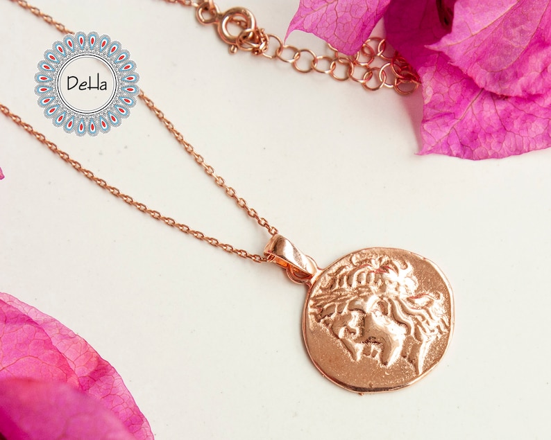 Pode incluir: Colar em ouro rosa com um pendente circular com um relevo cl&aacute;ssico. A corrente e o pendente s&atilde;o exibidos sobre um fundo neutro, com elementos florais cor-de-rosa. Uma joia delicada.
