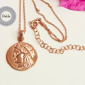 Pode incluir: Colar em ouro rosa com um pingente em forma de moeda que retrata um perfil cl&aacute;ssico. O pingente tem uma superf&iacute;cie texturizada. A corrente &eacute; delicada e ajust&aacute;vel. O logotipo Della &eacute; vis&iacute;vel no canto superior esquerdo. Uma joia.