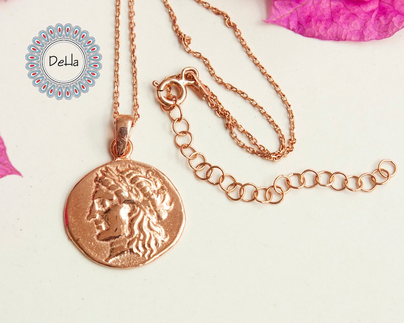 Pode incluir: Colar em ouro rosa com um pingente em forma de moeda que retrata um perfil cl&aacute;ssico. O pingente tem uma superf&iacute;cie texturizada. A corrente &eacute; delicada e ajust&aacute;vel. O logotipo Della &eacute; vis&iacute;vel no canto superior esquerdo. Uma joia.