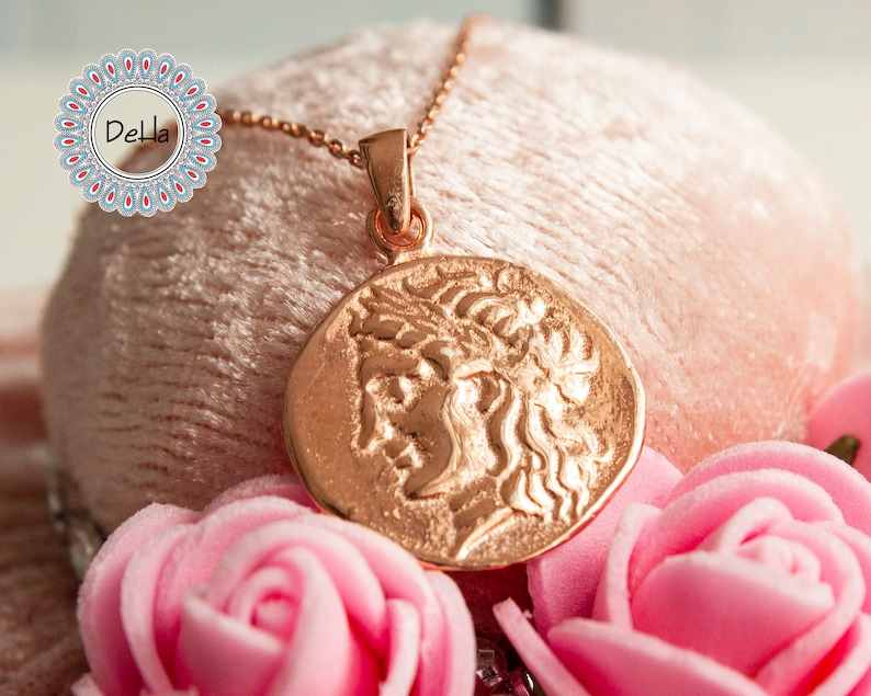 Pode incluir: Um colar em ouro rosa com um pingente redondo que apresenta um perfil detalhado. O pingente est&aacute; pendurado em uma corrente fina. A imagem inclui rosas cor-de-rosa e um fundo rosa suave.