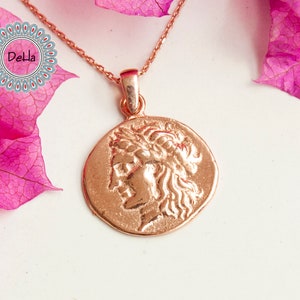 Pode incluir: Colar com pingente em ouro rosa com um perfil detalhado de uma pessoa com uma coroa de louros. O pingente redondo pende de uma corrente delicada. O fundo inclui flores rosa e uma superf&iacute;cie branca.