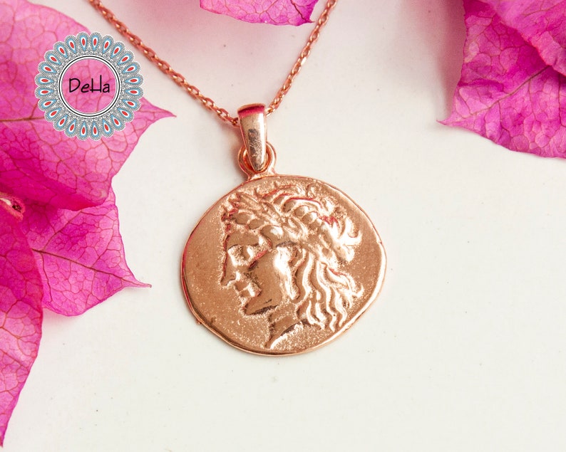 Pode incluir: Colar com pingente em ouro rosa com um perfil detalhado de uma pessoa com uma coroa de louros. O pingente redondo pende de uma corrente delicada. O fundo inclui flores rosa e uma superf&iacute;cie branca.