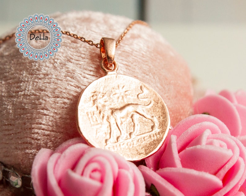 Pode incluir: Um colar com pingente em ouro rosa com um charme redondo em forma de moeda com um design em relevo de uma figura e texto. O colar &eacute; exibido em uma superf&iacute;cie rosa com rosas cor-de-rosa em primeiro plano. O logotipo Della &eacute; vis&iacute;vel.