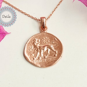 Pode incluir: Colar em tom de ouro rosa com um pingente redondo que apresenta um le&atilde;o e texto em relevo. O pingente est&aacute; suspenso por uma corrente delicada. O logotipo Della &eacute; vis&iacute;vel no canto superior esquerdo, e flores cor-de-rosa est&atilde;o no fundo.
