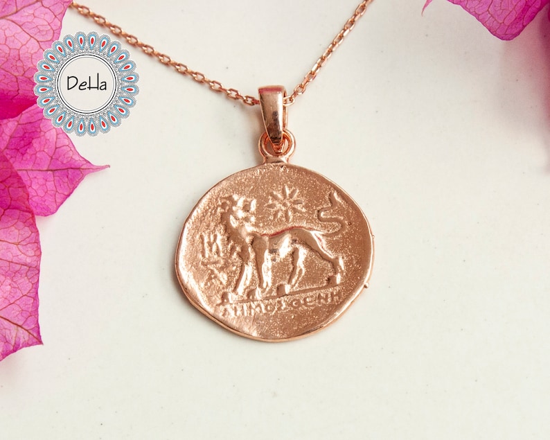 Pode incluir: Colar em tom de ouro rosa com um pingente redondo que apresenta um le&atilde;o e texto em relevo. O pingente est&aacute; suspenso por uma corrente delicada. O logotipo Della &eacute; vis&iacute;vel no canto superior esquerdo, e flores cor-de-rosa est&atilde;o no fundo.