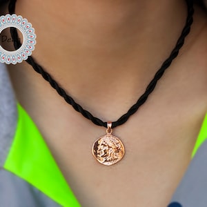 Pode incluir: Um colar com pingente em ouro rosa com um pingente redondo em forma de moeda com um design em relevo detalhado. O pingente pende de um colar de cord&atilde;o tran&ccedil;ado preto. O colar &eacute; exibido em um fundo neutro.