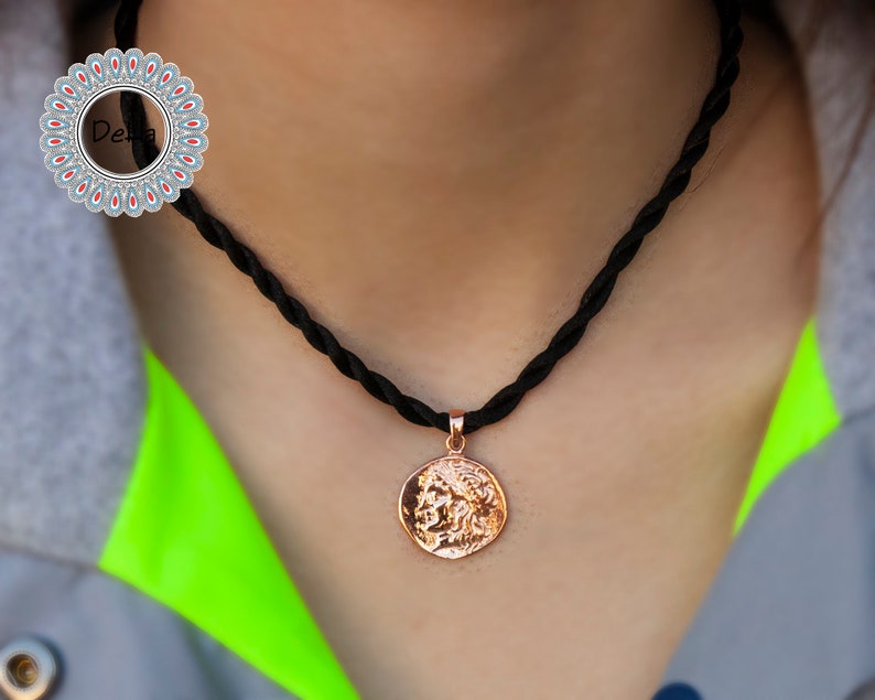 Pode incluir: Um colar com pingente em ouro rosa com um pingente redondo em forma de moeda com um design em relevo detalhado. O pingente pende de um colar de cord&atilde;o tran&ccedil;ado preto. O colar &eacute; exibido em um fundo neutro.
