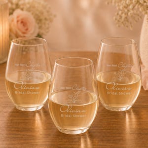 Op de afbeelding: Drie stamloze wijnglazen gevuld met een lichtgekleurde vloeistof. Elk glas is geëtst met de tekst "Her Next Chapter", een bloemenontwerp en "Olivia's Bridal Shower". De glazen staan op een houten oppervlak.