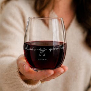 May include: A stemless wine glass filled with red wine. The glass has the text "olivia's godmother" and "EST 2026" etched on the front. The glass is held in a person's hand.