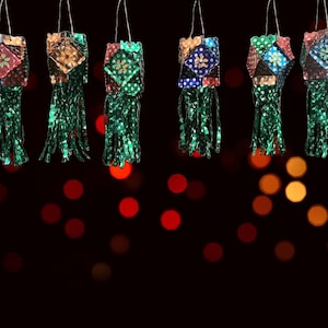 May include: Six colorful, hanging lanterns with square tops and green fringe. The lantern tops feature various patterns and colors, including red, blue, and gold. The background is a dark, blurred bokeh of red and orange lights.