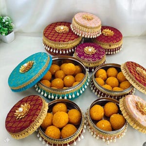 May include: A collection of round, decorative containers filled with golden-yellow sweets. The containers have ornate lids in various colors, including teal, burgundy, and pink, adorned with gold trim, tassels, and pearl accents. The lids are topped with a gold flower design.