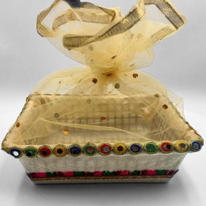 May include: A decorative gift basket with a rectangular shape. The basket is covered with a sheer, gold-colored fabric and adorned with gold sequins. The rim is decorated with colorful circular mirrors and trim.