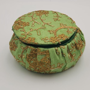 May include: A round, light green fabric pouch with a matching lid. The pouch and lid are adorned with gold-colored floral embroidery. The pouch has a gathered design around the base and a green zipper closure.