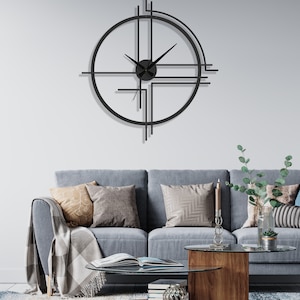 May include: A black metal wall clock with a geometric design. The clock has a round face with black hands and a black center. The clock is hanging on a white wall above a gray sofa.
