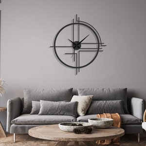 May include: A black metal geometric clock with a minimalist design hangs on a gray wall above a gray sofa with throw pillows and a wooden coffee table with a bowl of decorative items.