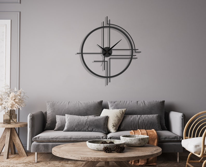 May include: A black metal geometric clock with a minimalist design hangs on a gray wall above a gray sofa with throw pillows and a wooden coffee table with a bowl of decorative items.