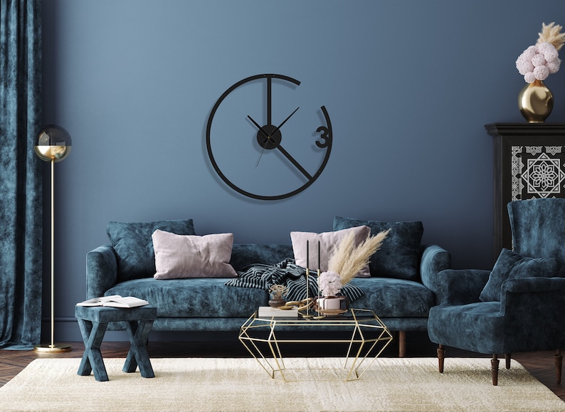 May include: A living room interior with a blue velvet sofa, a matching armchair, and a gold geometric coffee table. A black clock with a unique design hangs on the wall. The room is decorated with a vase of pink flowers and a floor lamp.