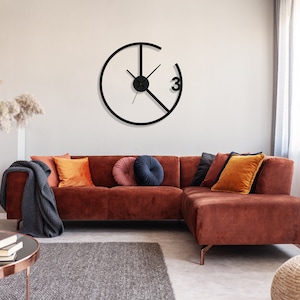 May include: A brown sectional sofa with a black throw blanket and orange and navy blue throw pillows. A black clock with a white face and black hands is hanging on the wall behind the sofa.