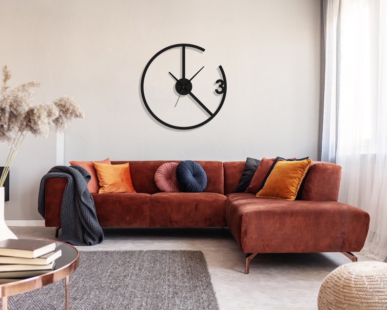 May include: A brown sectional sofa with a black throw blanket and orange and navy blue throw pillows. A black clock with a white face and black hands is hanging on the wall behind the sofa.