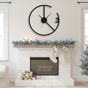 May include: A black metal clock with a circular design and Roman numeral markings. The clock is hanging on a white wall above a fireplace with a white brick surround. The fireplace has a black metal grate and a pile of logs in front of it. There are two white stockings hanging from the mantelpiece.
