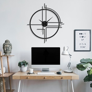 May include: A black metal clock with a geometric design hangs on a white wall above a wooden desk. The clock has a circular face with black hands and a black ring around the edge. The desk has a computer monitor, a lamp, a plant, and a cup on it. The wall also has a framed print with the text "Don't grow up, it's a trap" on it.