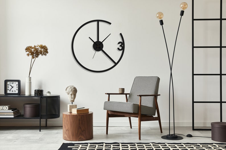 May include: A black metal wall clock with a unique design featuring a circle with a smaller circle inside and a diagonal line intersecting the circle. The clock has black hands and the number "3" is visible on the clock face.
