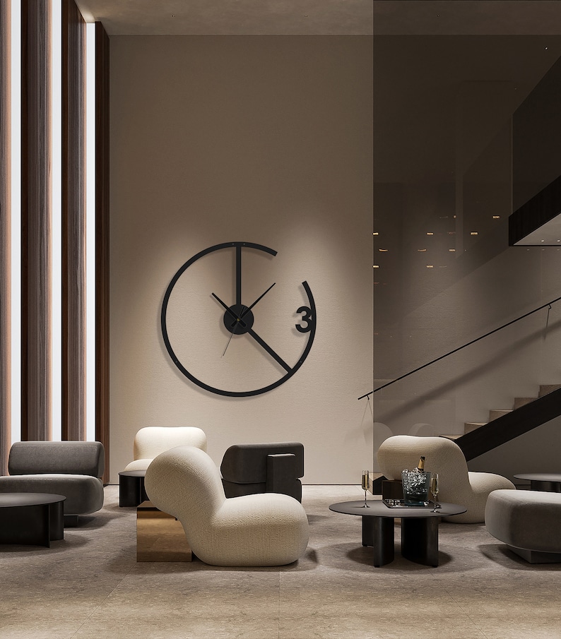 May include: A modern black wall clock with a circular design and a single Roman numeral "3" on the face. The clock is hanging on a beige wall in a living room setting with two white armchairs and a black coffee table.
