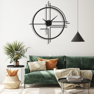 May include: A black metal wall clock with a geometric design. The clock is hanging on a white wall above a green velvet sofa with throw pillows and a blanket.