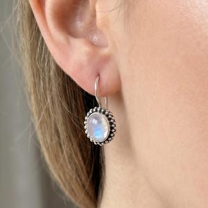 Pode incluir: Um brinco de prata com um cabochão redondo e iridescente de pedra da lua. A pedra é rodeada por uma borda de prata com contas. O brinco é do estilo pendente, com um gancho simples.