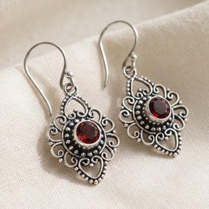 May include: A pair of silver drop earrings featuring a round, deep red gemstone set in the centre of an intricate, filigree-style metal frame. The earrings hang from simple silver hooks and display a detailed, textured pattern around the stone.