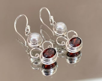 May include: Silver earrings with a drop design. Each earring features a lustrous, round pearl and a deep red oval gemstone. The earrings have a hook closure and are displayed on a reflective surface.