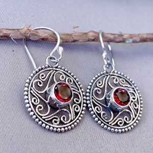 May include: A pair of silver-coloured drop earrings. Each earring has a circular, ornate design with a central, round, deep red gemstone. The earrings feature a detailed, openwork pattern and a beaded border. The earrings are suspended from a simple hook.
