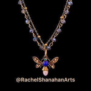 May include: A delicate gold chain necklace with a blue and green jeweled bee pendant. The bee has a pearl body and clear crystal wings. The chain is adorned with iridescent blue beads.