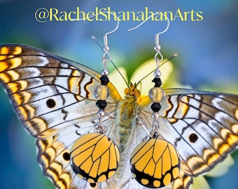 May include: A pair of dangle earrings with black and clear beads and yellow butterfly-shaped charms. The earrings are photographed against a butterfly with white and yellow wings.