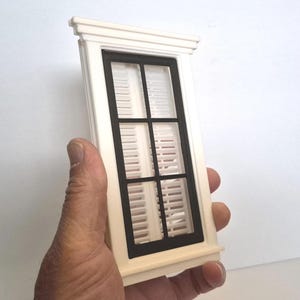 Könnte beinhalten: Ein Miniatur-Fensterrahmen mit weißem Außenrahmen und dekorativen Zierleisten oben und unten. Der innere Bereich ist schwarz mit einem sechsfeldrigen Gitterdesign, und hinter dem schwarzen Fensterrahmen sind weiße Lamellenläden zu sehen.