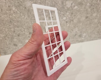 May include: A white, rectangular window frame with a grid pattern, held in a hand. The frame is made of a solid, opaque material and features a series of vertical and horizontal lines creating multiple small rectangular openings. The window frame is a miniature architectural element.