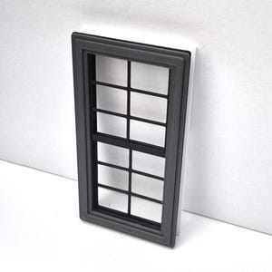 May include: A miniature window with a dark gray frame and black grid pattern. The window is set against a white background, creating a contrast. The design is simple and geometric, suitable for dollhouses or model projects.