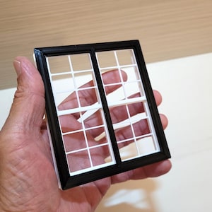 May include: A small, black and white window frame with a grid pattern. The window is held in a hand, highlighting its size and intricate design. The frame is black, and the window panes are white.