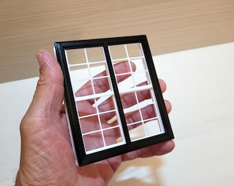 May include: A small, black and white window frame with a grid pattern. The window is held in a hand, highlighting its size and intricate design. The frame is black, and the window panes are white.