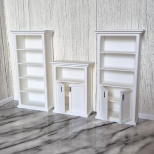 May include: Three white miniature cabinets arranged in a row. Two tall units feature five open shelves each, while the central shorter unit includes an upper open shelf and a lower cabinet section with two small doors featuring black vertical handles.