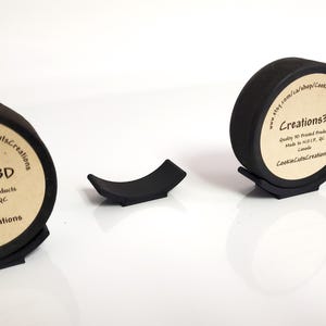 May include: Black 3D-printed hockey puck display stands. The stands are designed to hold hockey pucks upright. The pucks have a label with the text "Creations3D". The stands are made of black plastic and are designed to be durable.