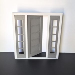 May include: A papercraft model of a modern front door in grey and white. The door is ajar, showing a white interior. The sidelights feature rectangular window cutouts. The design is clean and contemporary, suitable for a modern home.